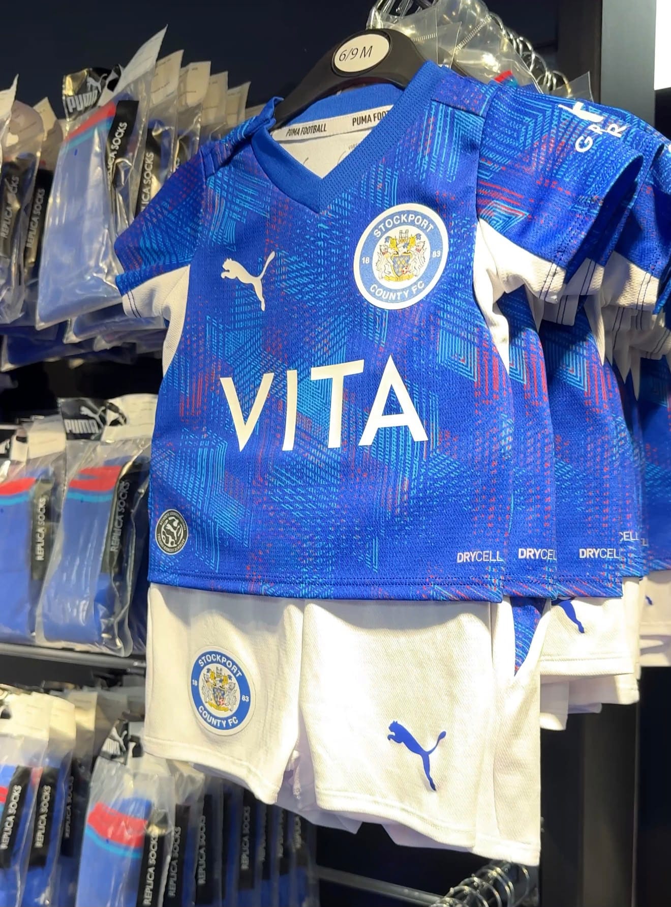 On a hanger in the shop, a baby’s blue patterned shirt with the Stockport County crest, and its white shorts